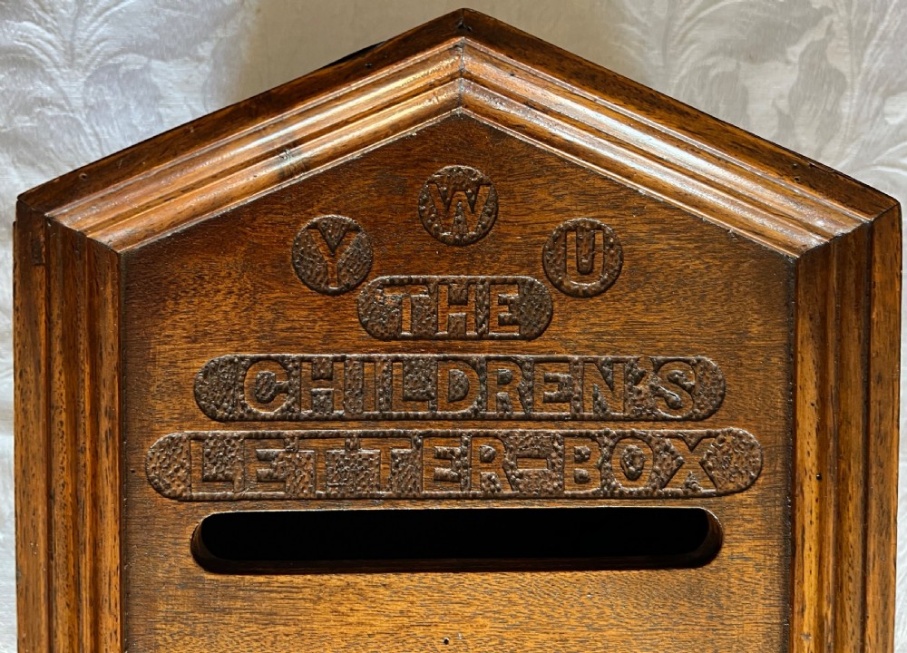 A wall-mounted carved oak letter box with a pointed top, horizontal aperture slot, side metal catch, and carved emblems on the front panel. View IV