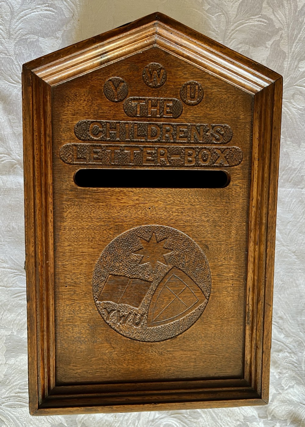 A wall-mounted carved oak letter box with a pointed top, horizontal aperture slot, side metal catch, and carved emblems on the front panel. View IX