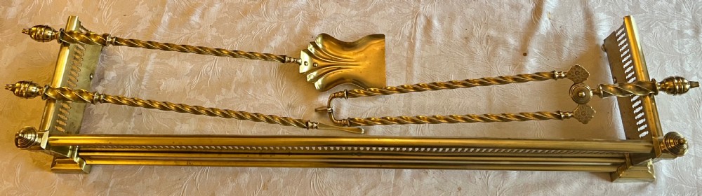A Victorian polished brass fireplace fender featuring integrated rail ends and a set of three matching barley twist brass fire irons including shovel, tongs, and poker.