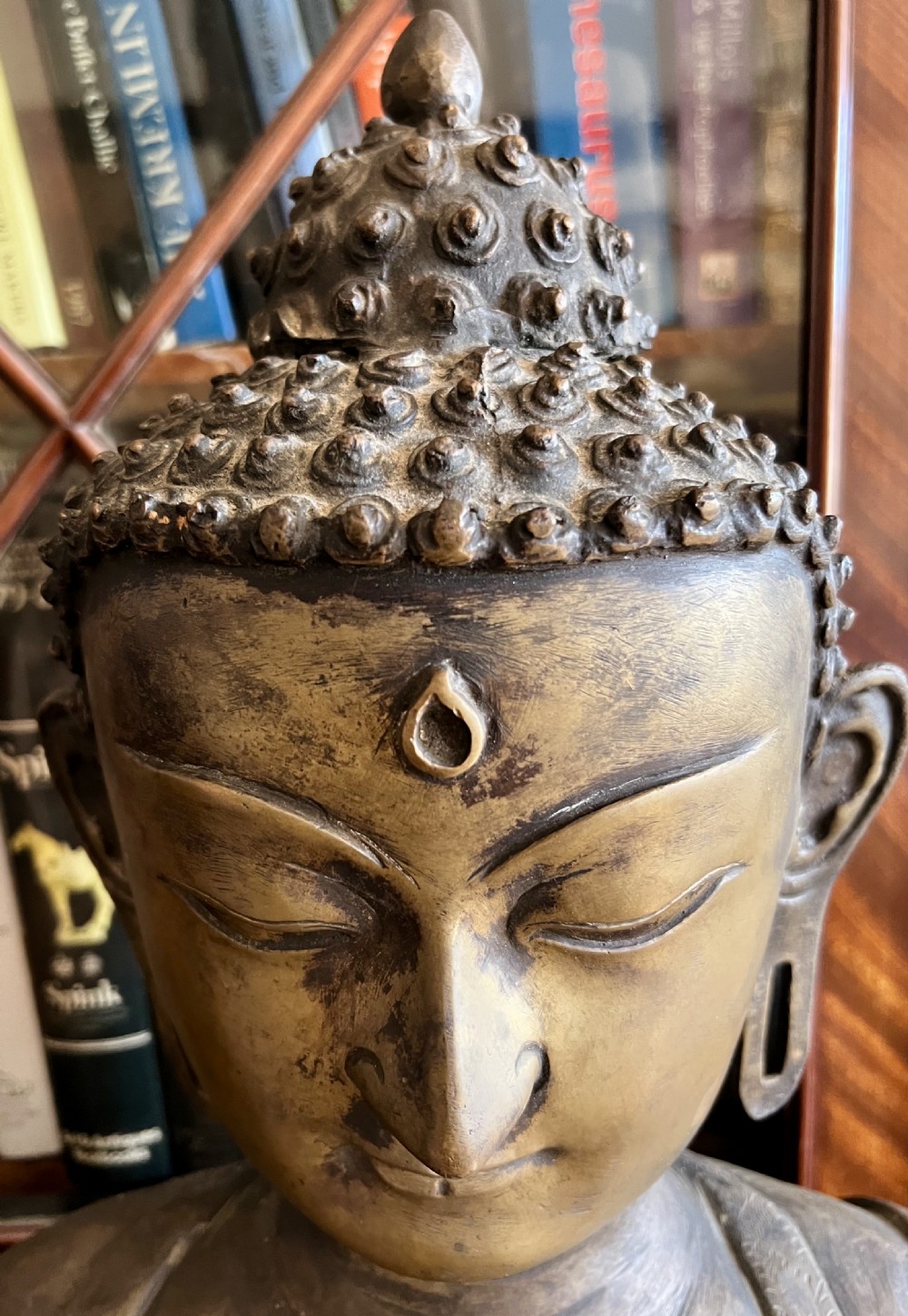 A cast bronze bust depicting a Buddha with downcast eyes, stylized hair curls, elongated earlobes, and draped robes. View III
