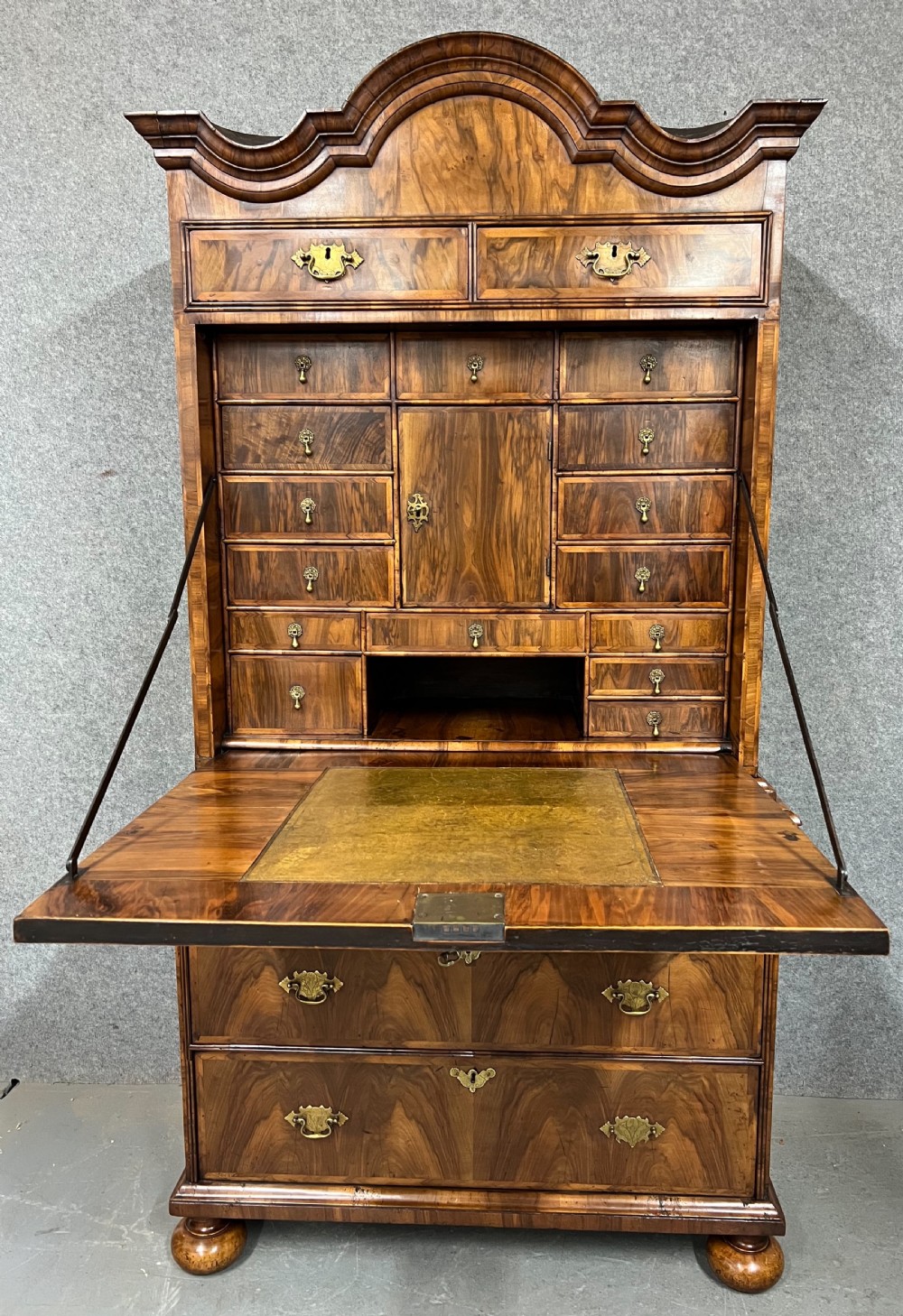 A William and Mary figured walnut escritoire featuring a double-dome arched pediment, a fall-front writing surface concealing a multi-drawer fitted interior, and a chest base on turned bun feet.