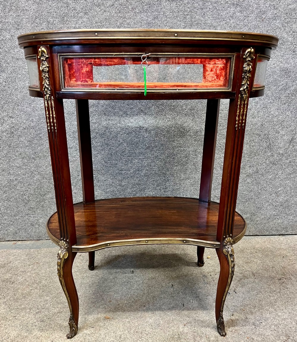 An oval French bijouterie table featuring mahogany wood framing, glass display panels lined with red fabric, gilt brass mounts, and fluted legs connected by a lower shelf.