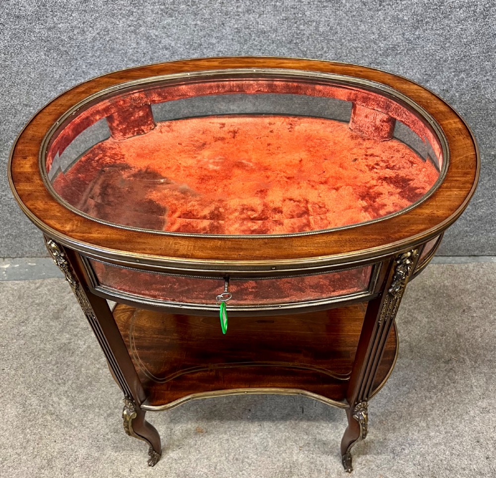 An oval French bijouterie table featuring mahogany wood framing, glass display panels lined with red fabric, gilt brass mounts, and fluted legs connected by a lower shelf. View II