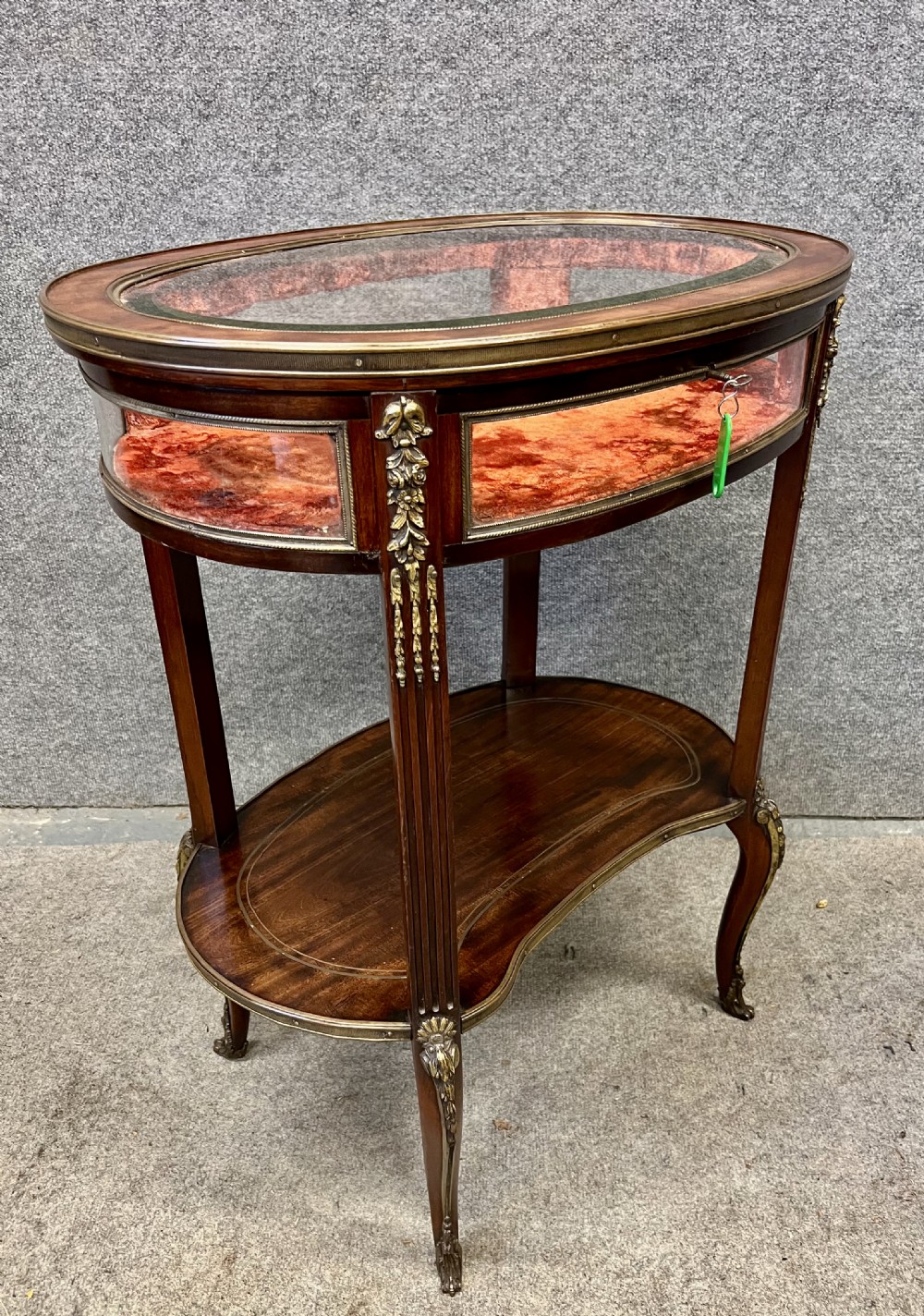 An oval French bijouterie table featuring mahogany wood framing, glass display panels lined with red fabric, gilt brass mounts, and fluted legs connected by a lower shelf. View IV
