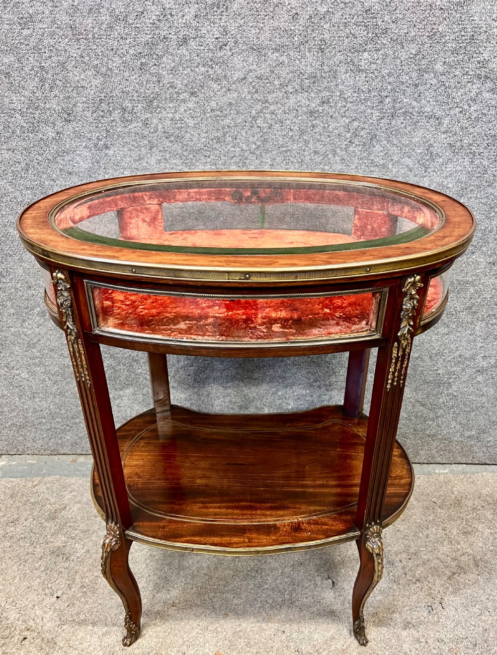 An oval French bijouterie table featuring mahogany wood framing, glass display panels lined with red fabric, gilt brass mounts, and fluted legs connected by a lower shelf. View VI