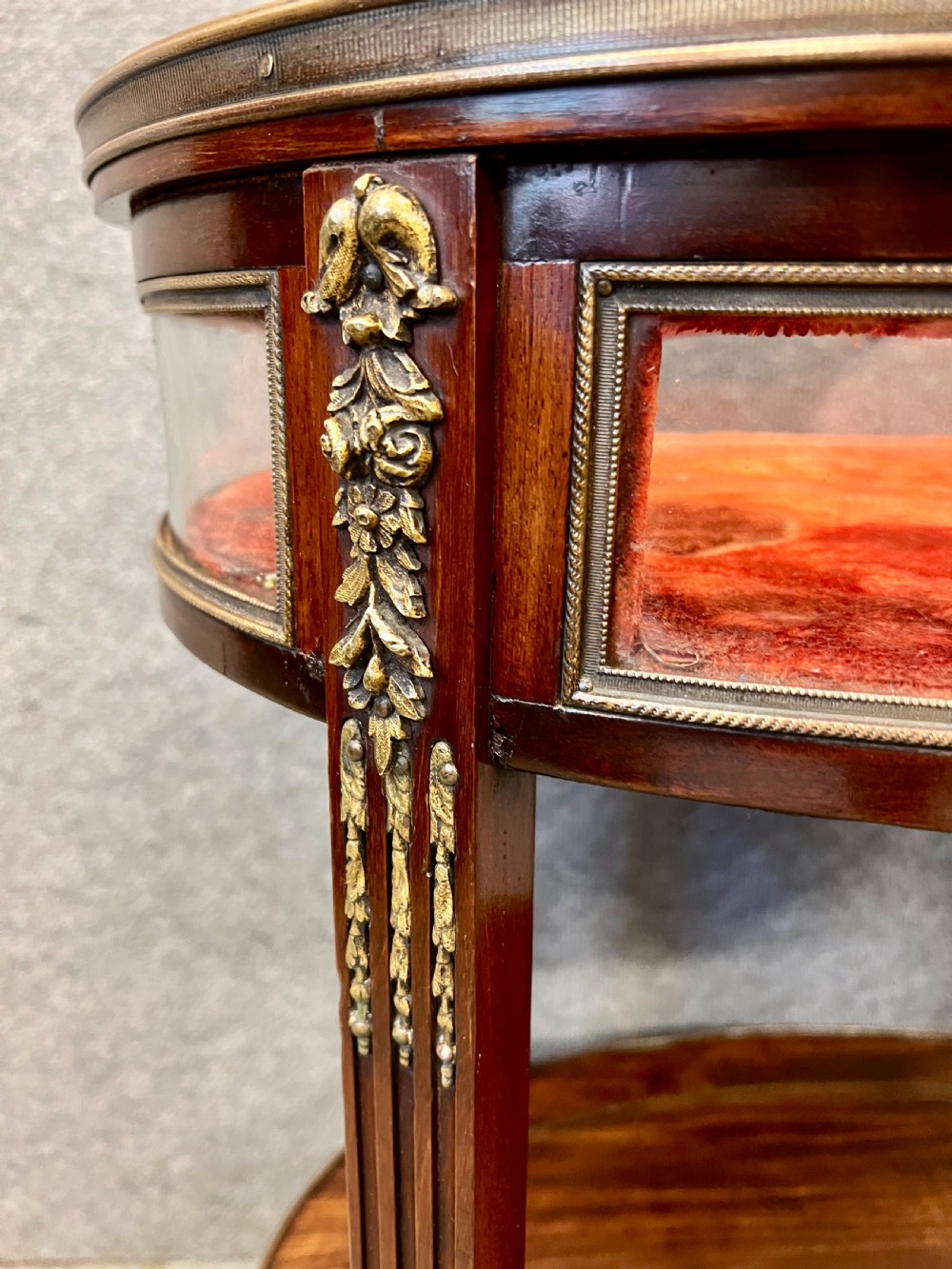 An oval French bijouterie table featuring mahogany wood framing, glass display panels lined with red fabric, gilt brass mounts, and fluted legs connected by a lower shelf. View IX