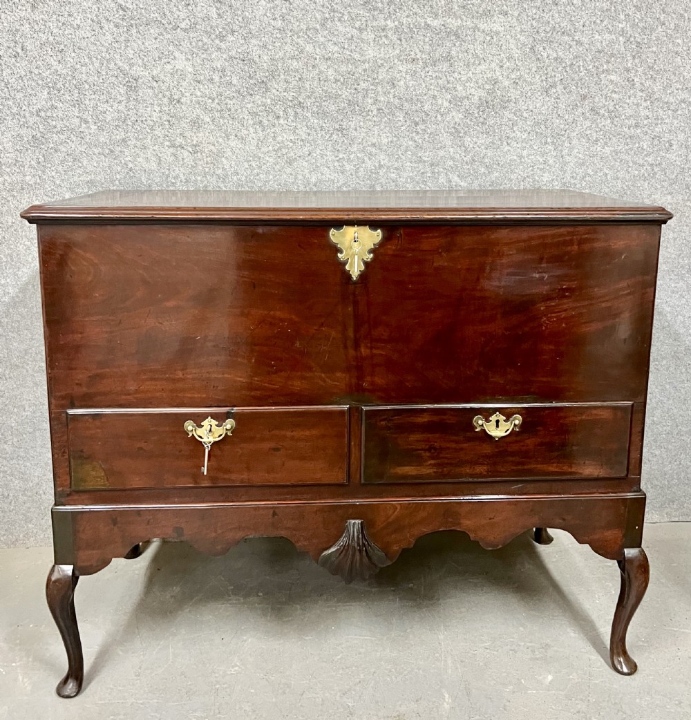 A George II mahogany chest on stand featuring a hinged top, two lower drawers with brass keyplates, a central carved shell apron, and cabriole legs.