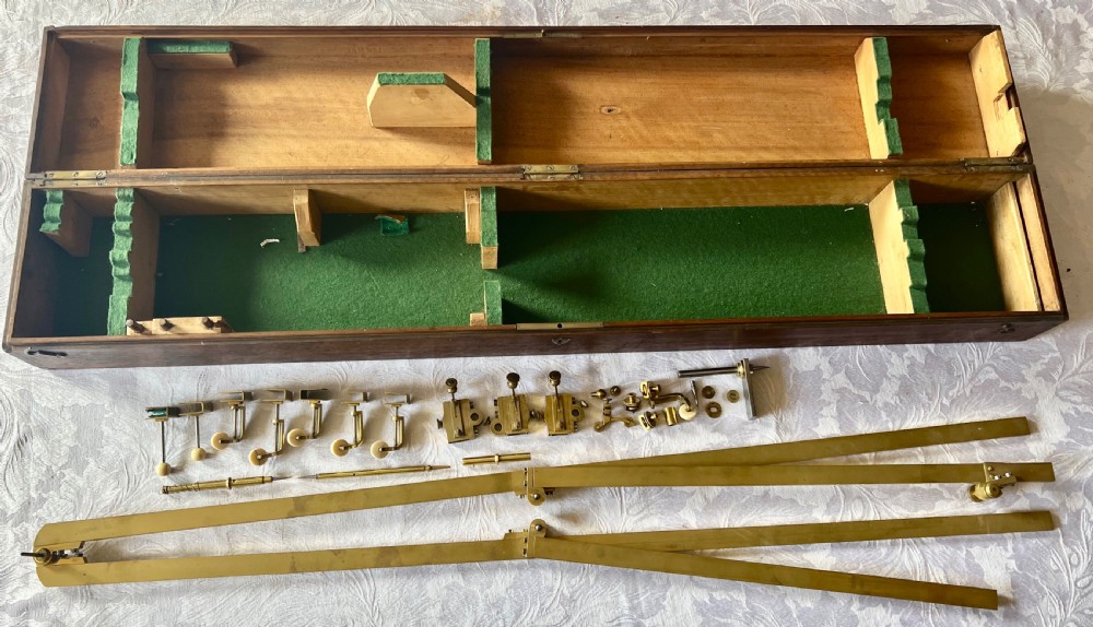 An antique brass pantograph with various small attachments laid out alongside its open fitted mahogany case with green felt lining.
