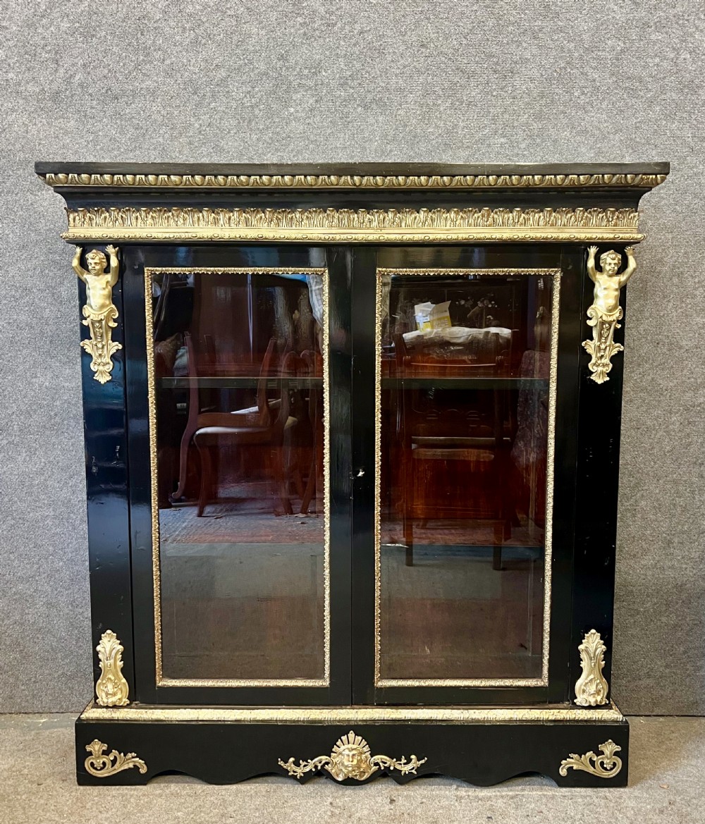 An ebonised two-door display cabinet featuring glass panels, detailed gilt bronze ormolu mounts, cherub term figures, and ornamental friezes.