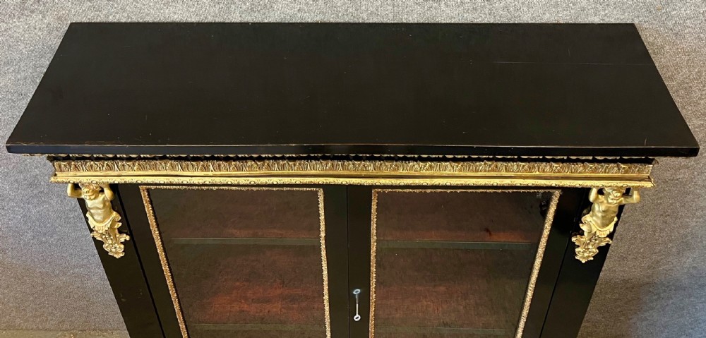 An ebonised two-door display cabinet featuring glass panels, detailed gilt bronze ormolu mounts, cherub term figures, and ornamental friezes. View IV
