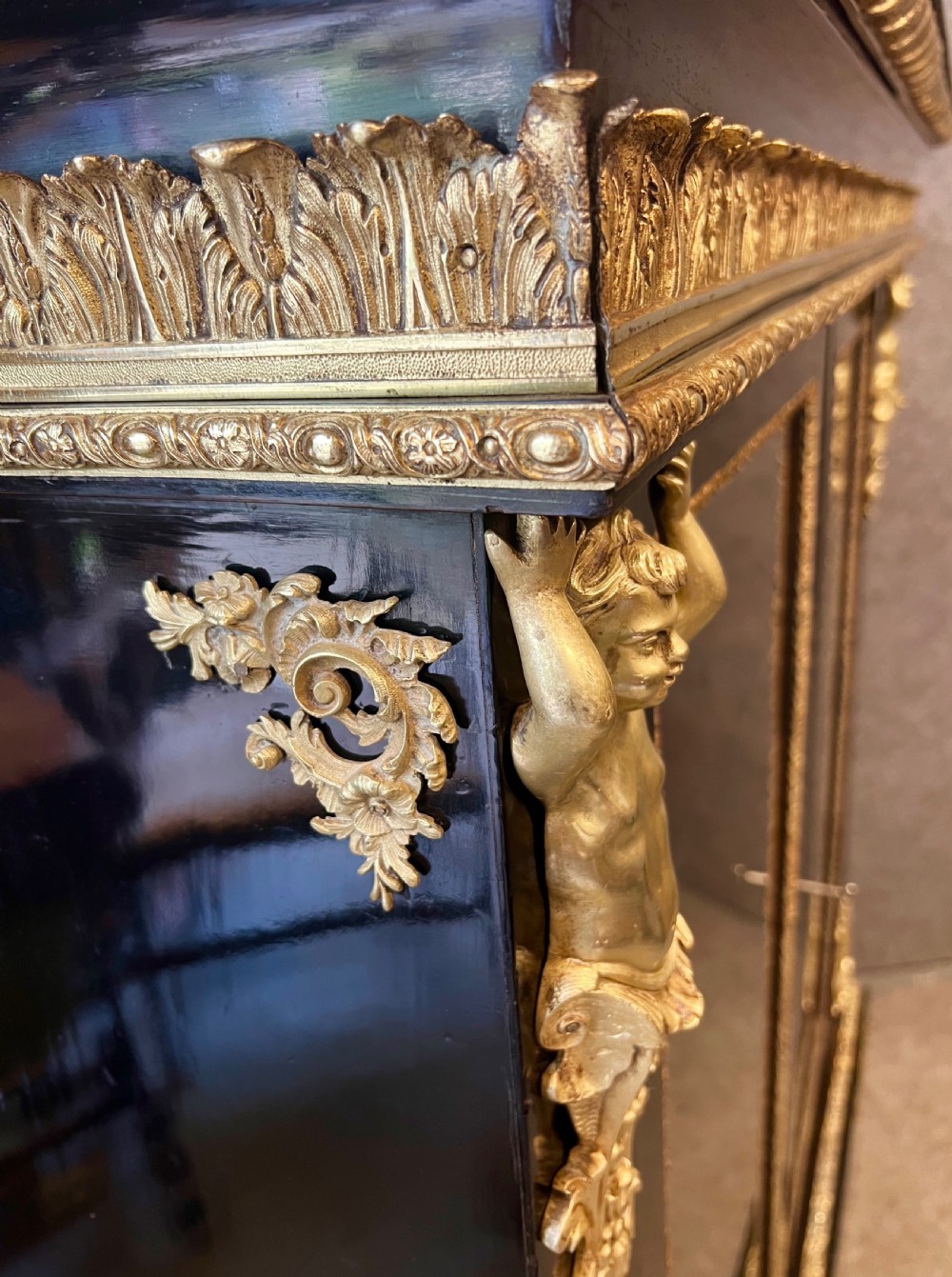 An ebonised two-door display cabinet featuring glass panels, detailed gilt bronze ormolu mounts, cherub term figures, and ornamental friezes. View VIII