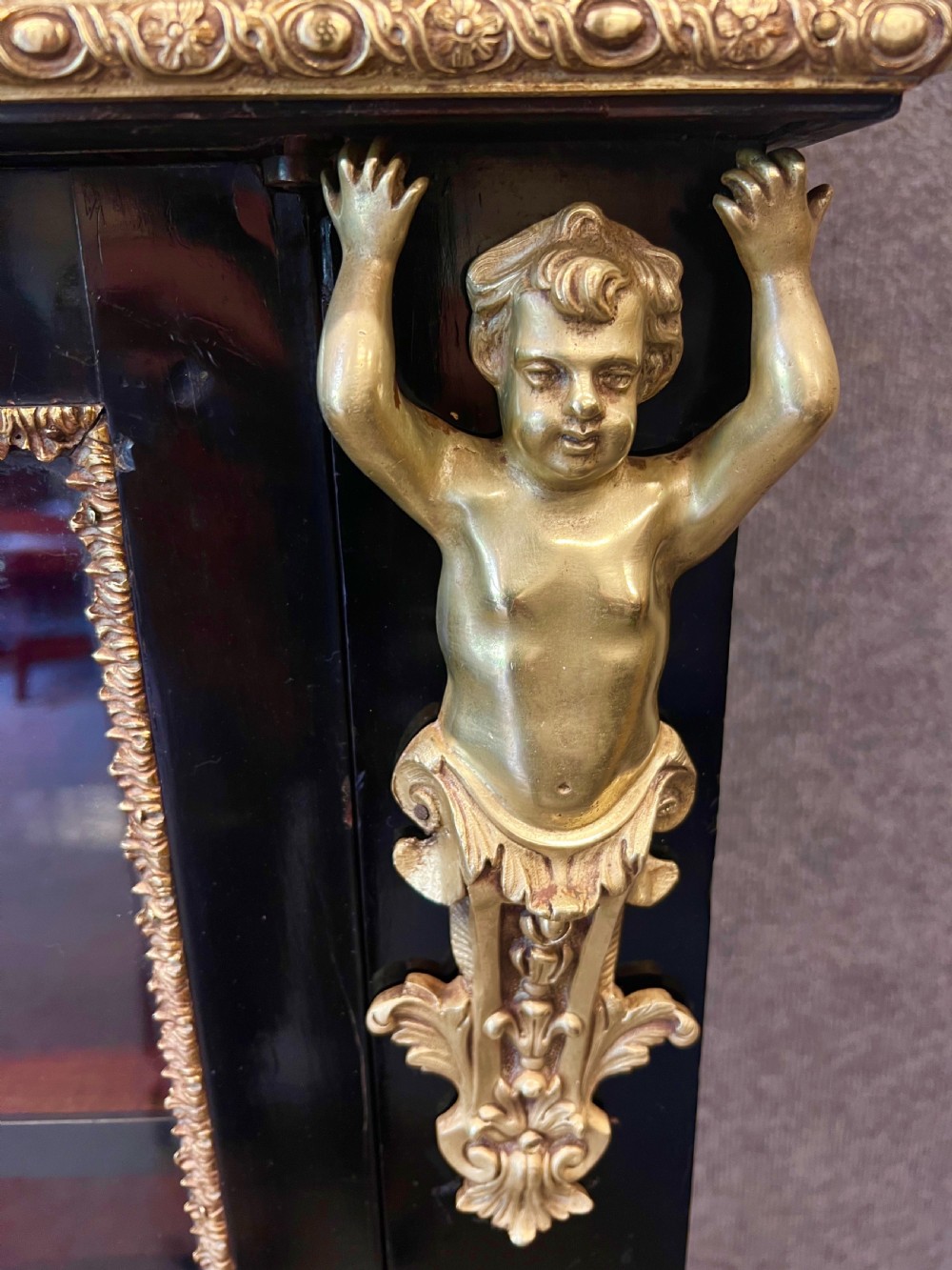 An ebonised two-door display cabinet featuring glass panels, detailed gilt bronze ormolu mounts, cherub term figures, and ornamental friezes. View XIII