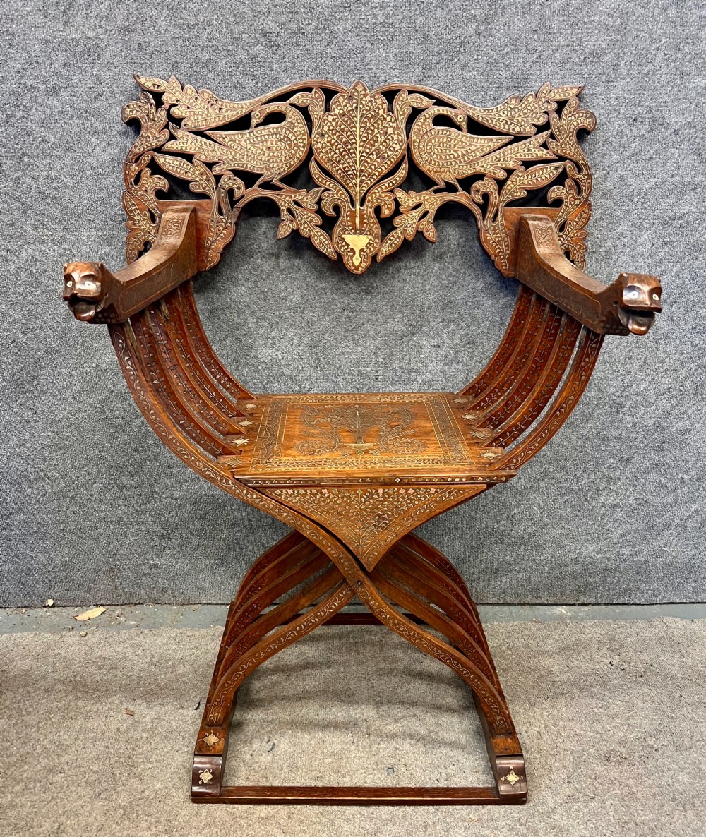 An Anglo-Indian X-frame Savonarola armchair crafted from dark carved wood featuring intricate brass inlay depicting peacocks and foliage with carved animal head armrests.