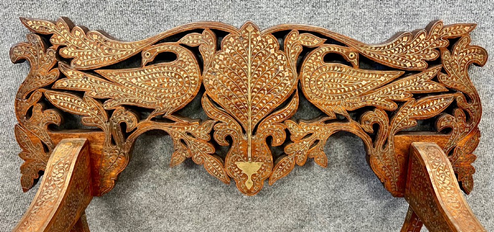 An Anglo-Indian X-frame Savonarola armchair crafted from dark carved wood featuring intricate brass inlay depicting peacocks and foliage with carved animal head armrests. View III