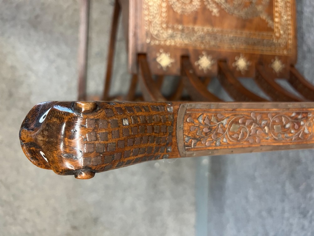 An Anglo-Indian X-frame Savonarola armchair crafted from dark carved wood featuring intricate brass inlay depicting peacocks and foliage with carved animal head armrests. View VI