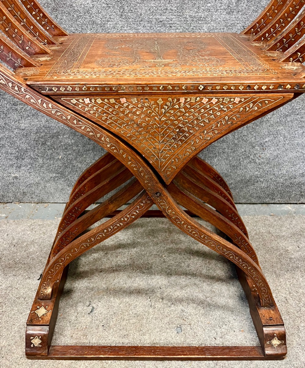 An Anglo-Indian X-frame Savonarola armchair crafted from dark carved wood featuring intricate brass inlay depicting peacocks and foliage with carved animal head armrests. View VII