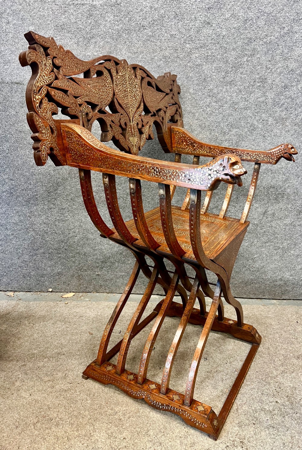 An Anglo-Indian X-frame Savonarola armchair crafted from dark carved wood featuring intricate brass inlay depicting peacocks and foliage with carved animal head armrests. View VIII