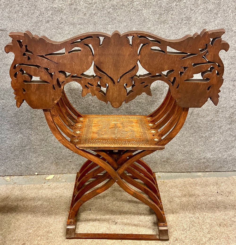 An Anglo-Indian X-frame Savonarola armchair crafted from dark carved wood featuring intricate brass inlay depicting peacocks and foliage with carved animal head armrests. View X