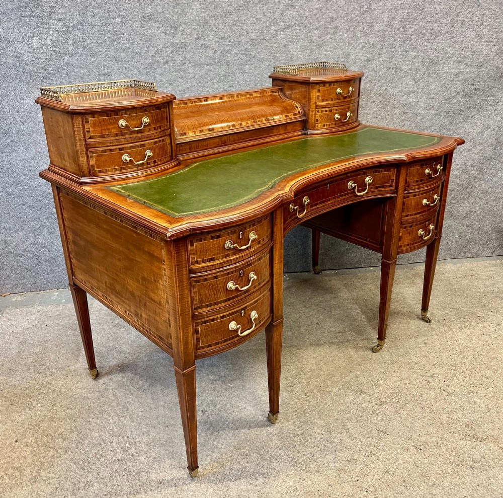 An antique mahogany Carlton House desk featuring satinwood inlay, a green leather writing surface, small raised drawers with brass gallery railings, and square tapered legs on castors.