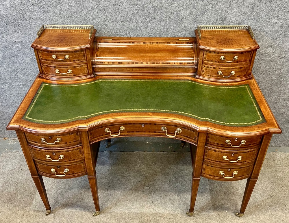 An antique mahogany Carlton House desk featuring satinwood inlay, a green leather writing surface, small raised drawers with brass gallery railings, and square tapered legs on castors. View II