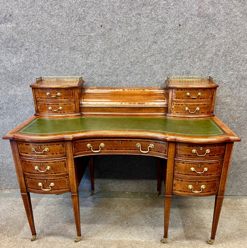 An antique mahogany Carlton House desk featuring satinwood inlay, a green leather writing surface, small raised drawers with brass gallery railings, and square tapered legs on castors. View III