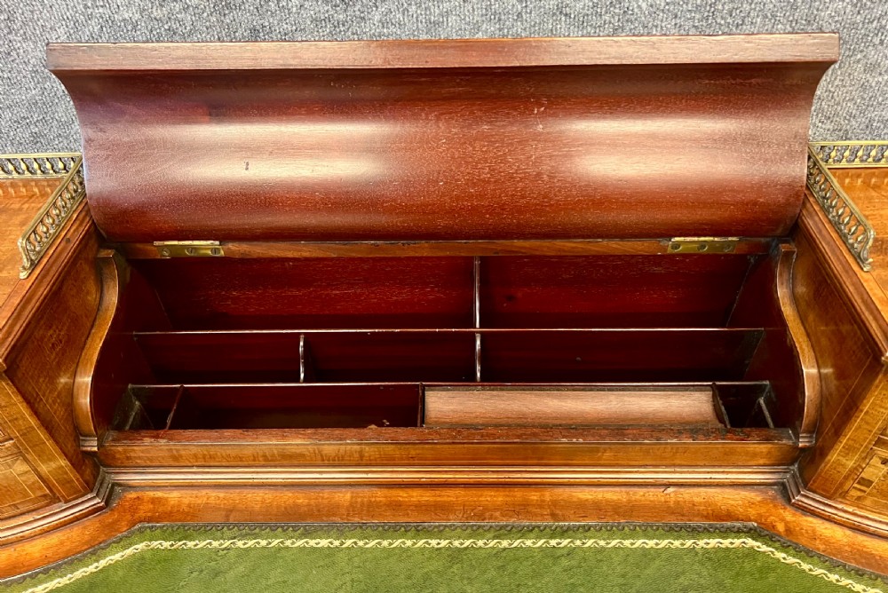 An antique mahogany Carlton House desk featuring satinwood inlay, a green leather writing surface, small raised drawers with brass gallery railings, and square tapered legs on castors. View V