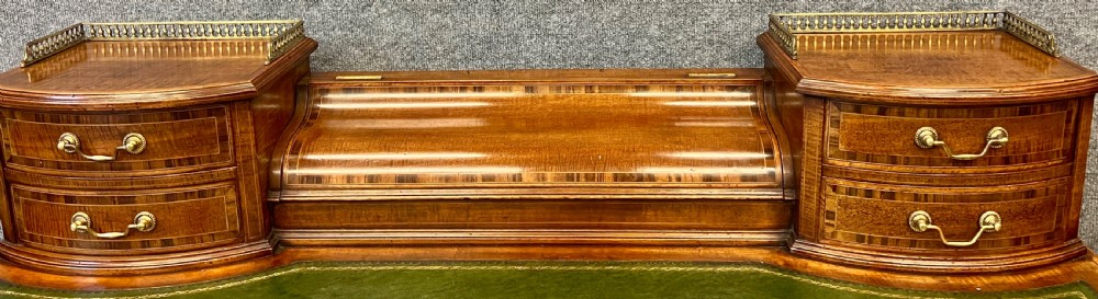 An antique mahogany Carlton House desk featuring satinwood inlay, a green leather writing surface, small raised drawers with brass gallery railings, and square tapered legs on castors. View VI