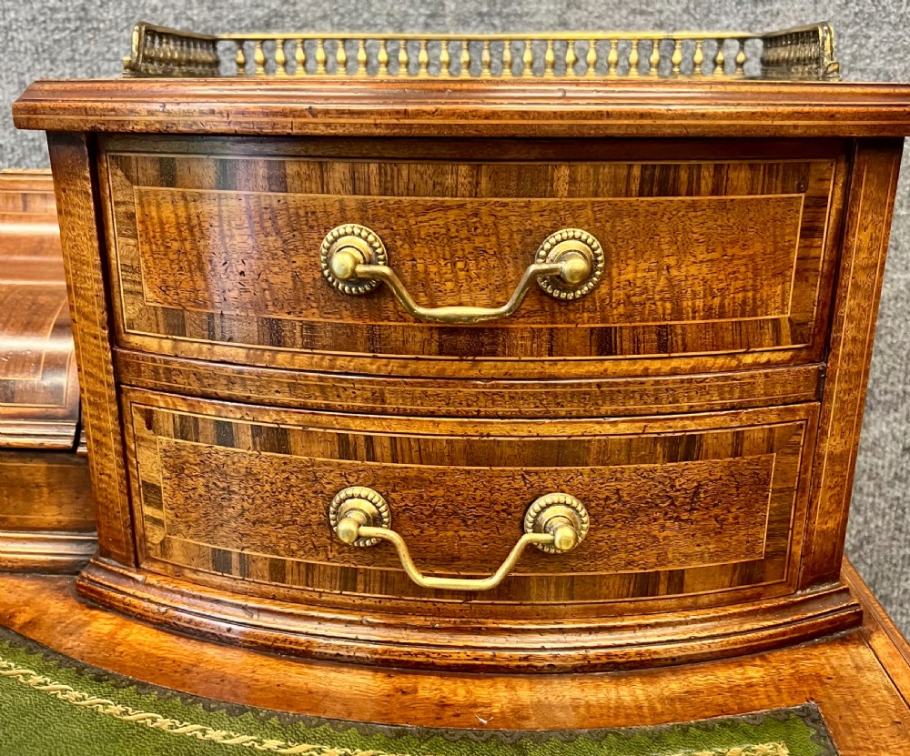 An antique mahogany Carlton House desk featuring satinwood inlay, a green leather writing surface, small raised drawers with brass gallery railings, and square tapered legs on castors. View VII