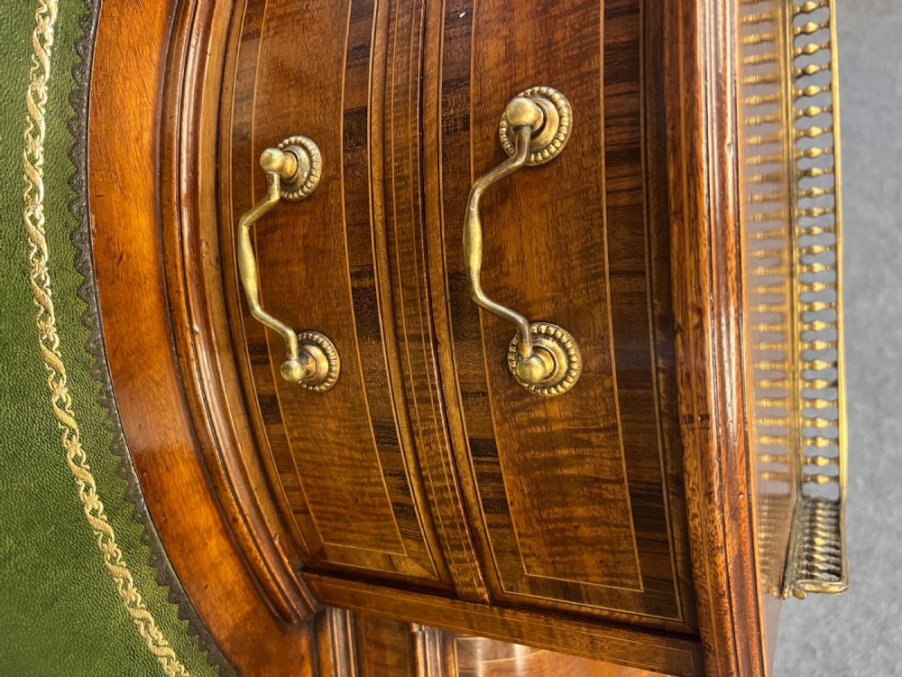 An antique mahogany Carlton House desk featuring satinwood inlay, a green leather writing surface, small raised drawers with brass gallery railings, and square tapered legs on castors. View VIII