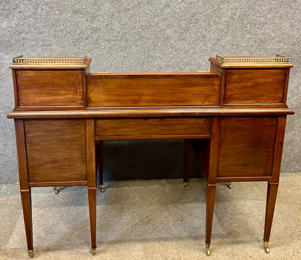 An antique mahogany Carlton House desk featuring satinwood inlay, a green leather writing surface, small raised drawers with brass gallery railings, and square tapered legs on castors. View IX