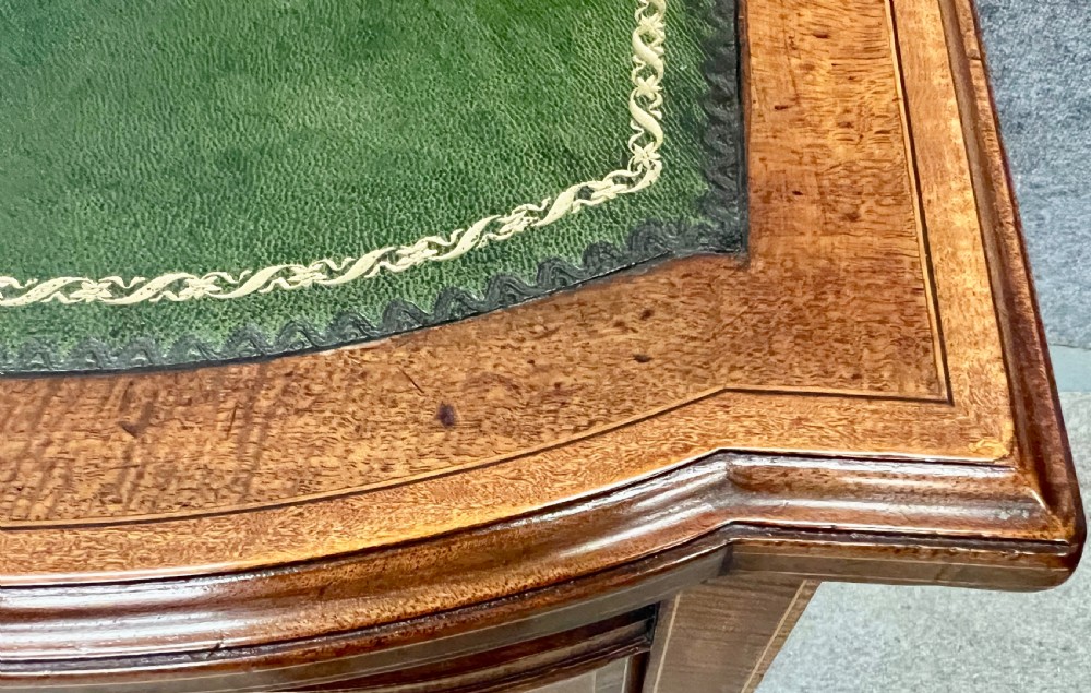 An antique mahogany Carlton House desk featuring satinwood inlay, a green leather writing surface, small raised drawers with brass gallery railings, and square tapered legs on castors. View XII