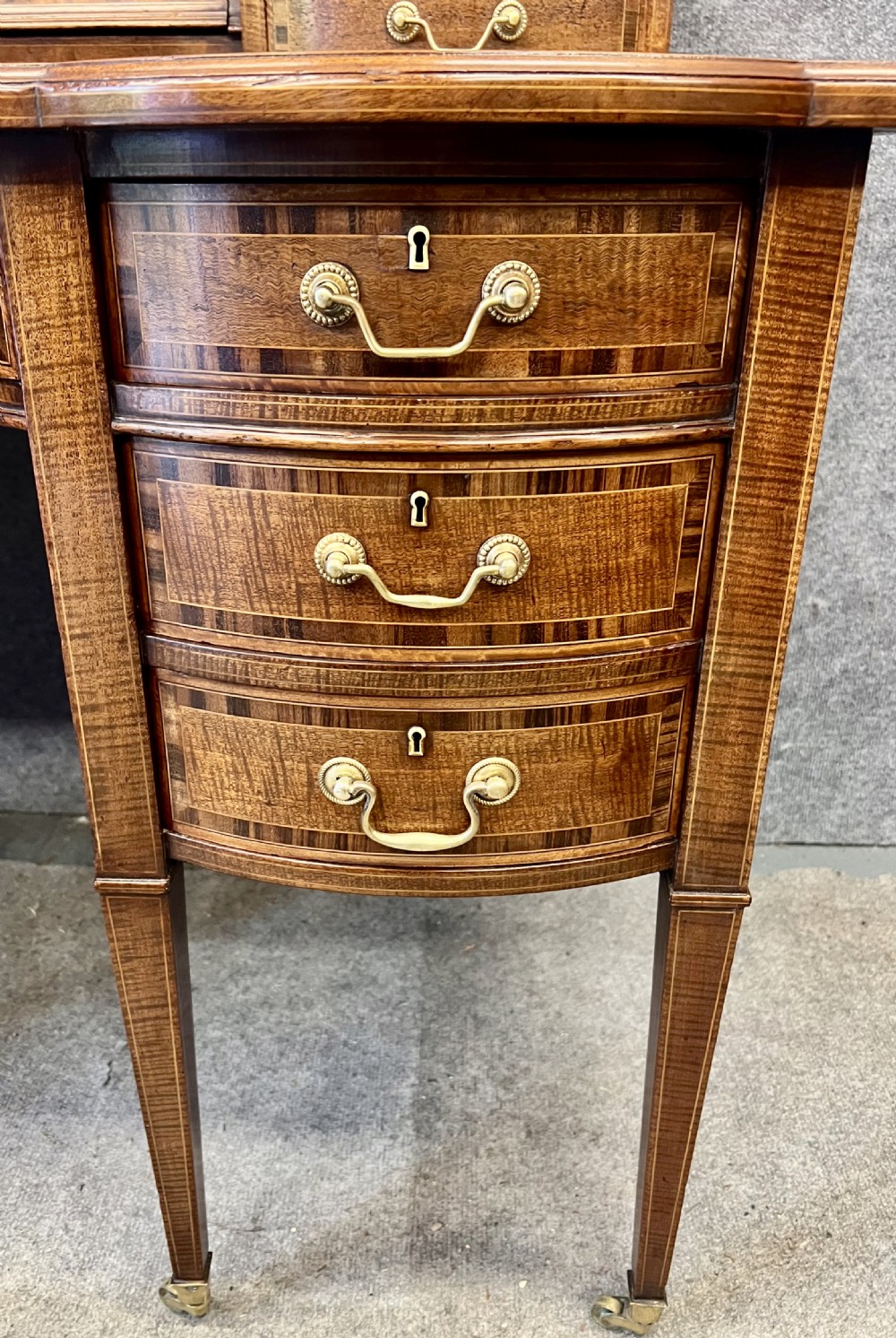 An antique mahogany Carlton House desk featuring satinwood inlay, a green leather writing surface, small raised drawers with brass gallery railings, and square tapered legs on castors. View XIII
