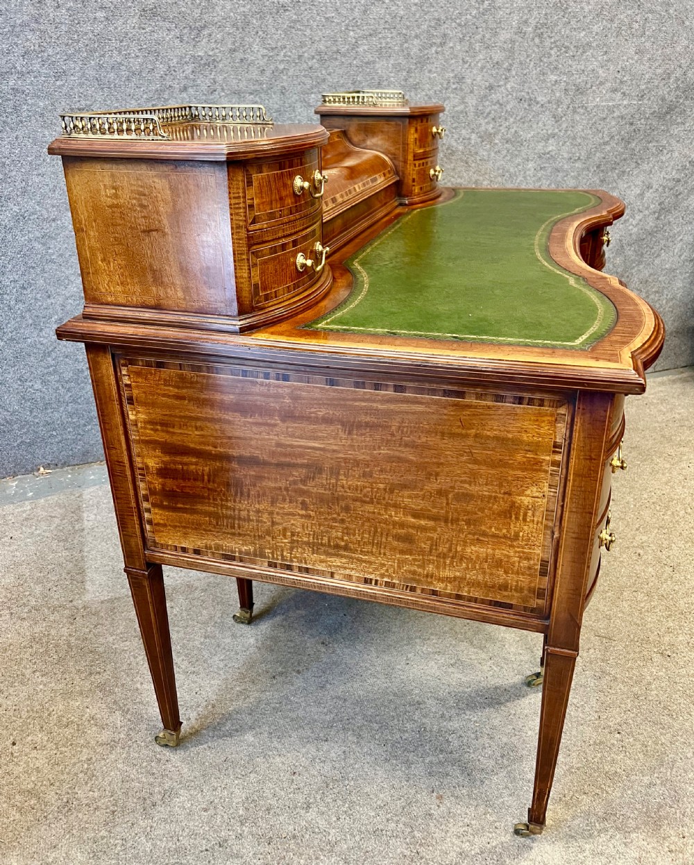 An antique mahogany Carlton House desk featuring satinwood inlay, a green leather writing surface, small raised drawers with brass gallery railings, and square tapered legs on castors. View XV