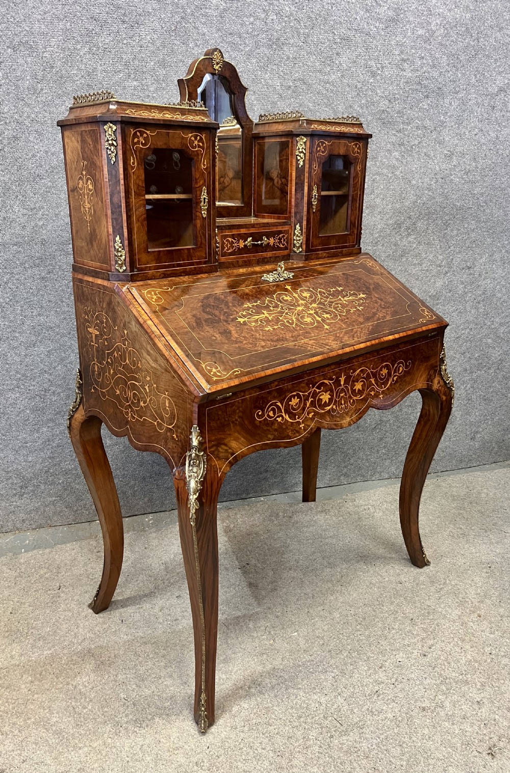 An antique Victorian inlaid walnut bureau de dame featuring a slant-front writing lid, floral marquetry, brass mounts, cabriole legs, and an upper cabinet with glazed doors.