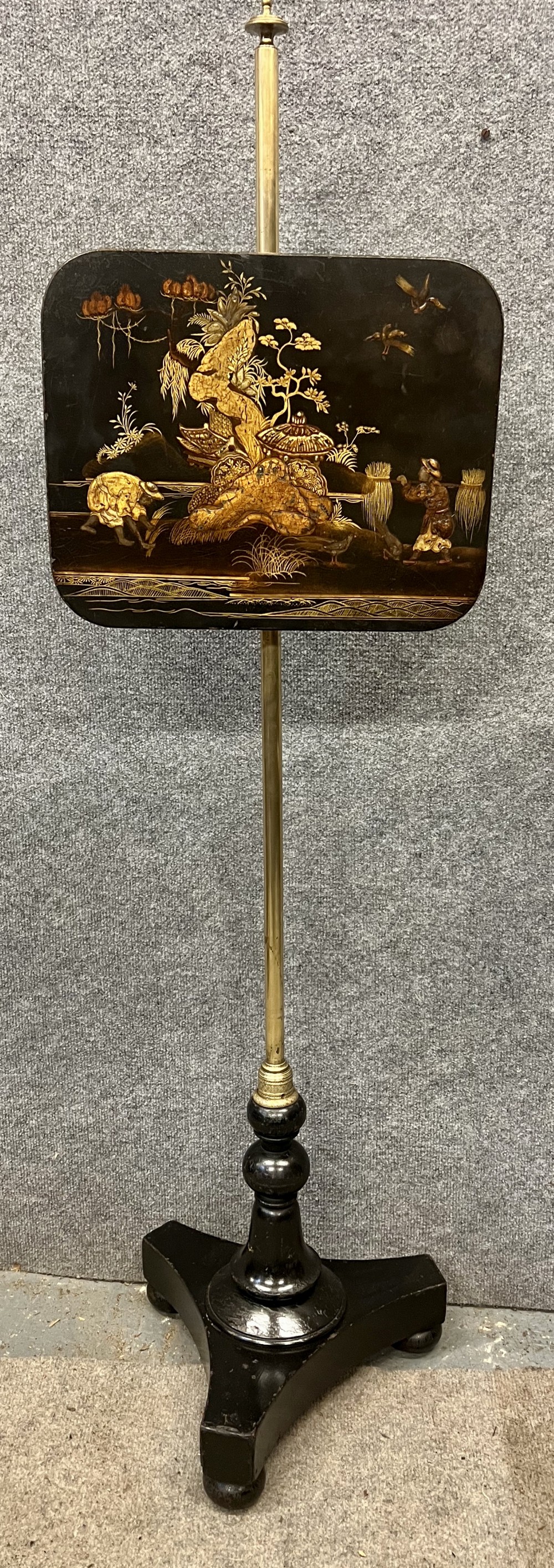 An antique Regency pole screen featuring a black lacquered rectangular panel decorated with gold Chinoiserie landscapes, supported by an adjustable brass rod on an ebonised tri-form base.