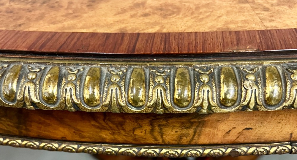 A Victorian burr walnut oval centre table featuring a crossbanded top with brass egg-and-dart moulding above a carved central column and four scrolled legs. View III