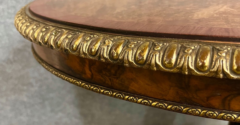 A Victorian burr walnut oval centre table featuring a crossbanded top with brass egg-and-dart moulding above a carved central column and four scrolled legs. View V