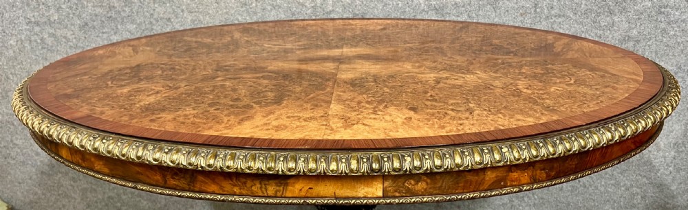 A Victorian burr walnut oval centre table featuring a crossbanded top with brass egg-and-dart moulding above a carved central column and four scrolled legs. View VI