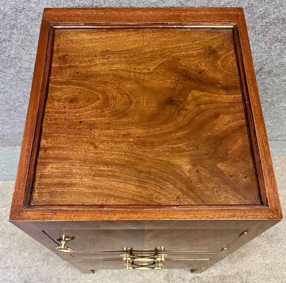 A George III mahogany bedside cabinet featuring a tray top, cupboard door, two drawers with brass swan-neck handles, and brass side carrying handles. View VI