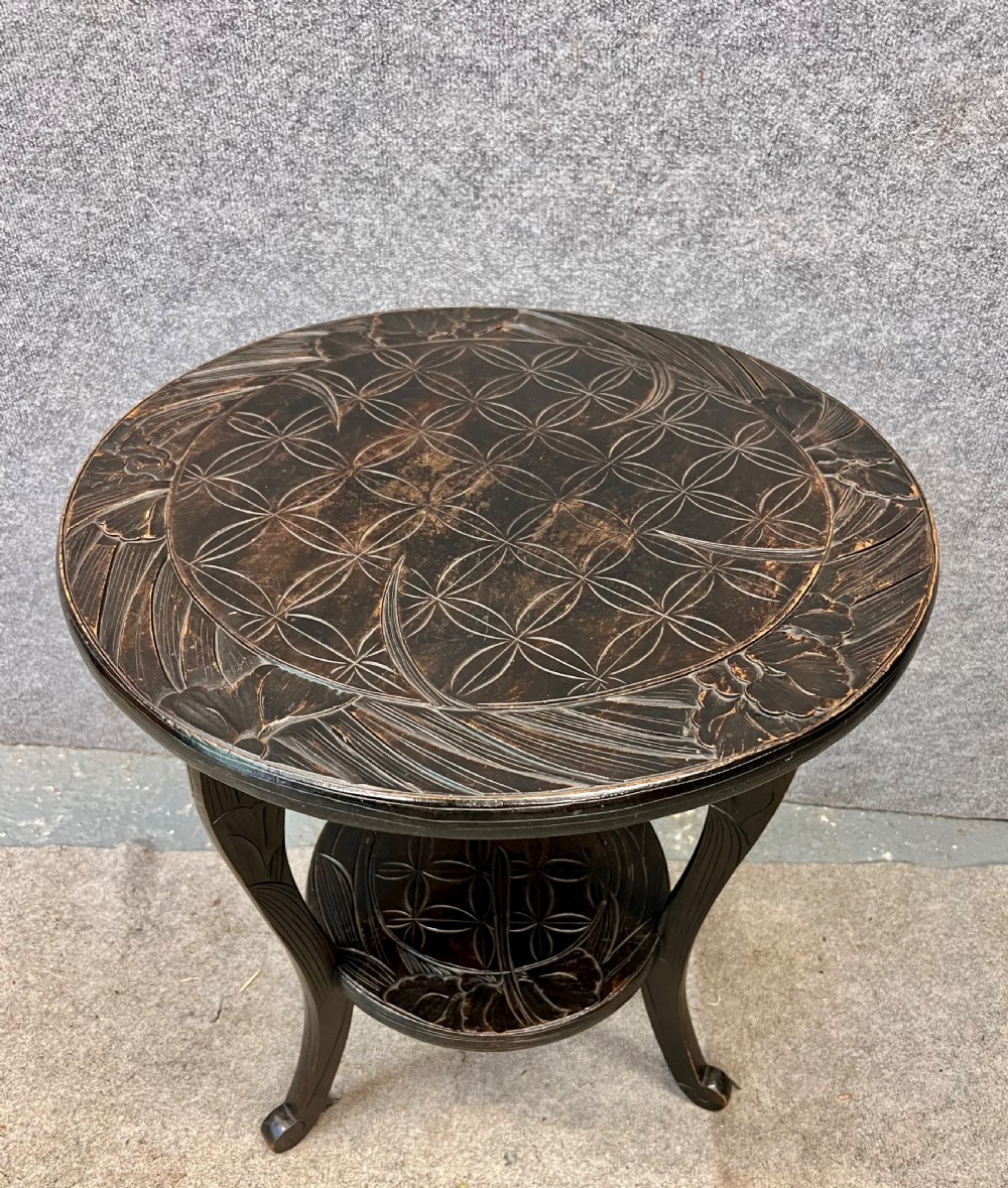 A circular dark ebonised wooden occasional table featuring a carved geometric top border with iris motifs, curved legs, and a small carved undertier shelf.