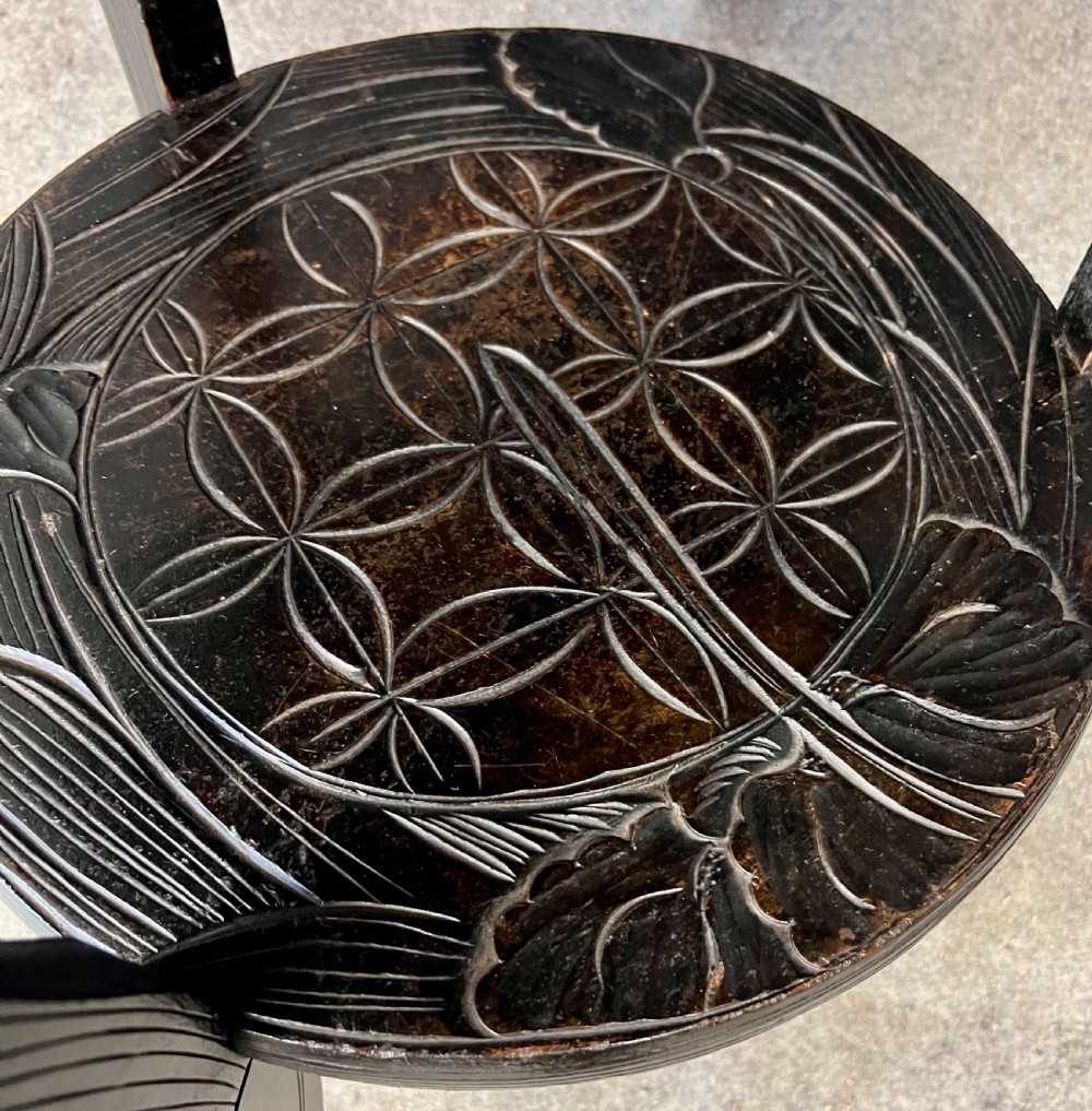 A circular dark ebonised wooden occasional table featuring a carved geometric top border with iris motifs, curved legs, and a small carved undertier shelf. View X