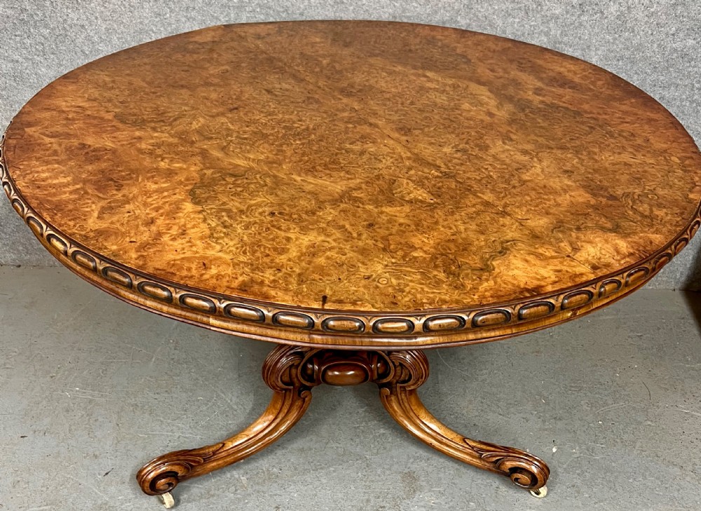 A circular Victorian centre table featuring a figured burr walnut top with carved egg-and-dart edge moulding, supported by a carved central pedestal with four splayed legs on ceramic castors.