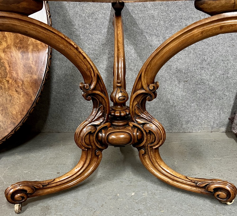 A circular Victorian centre table featuring a figured burr walnut top with carved egg-and-dart edge moulding, supported by a carved central pedestal with four splayed legs on ceramic castors. View IV