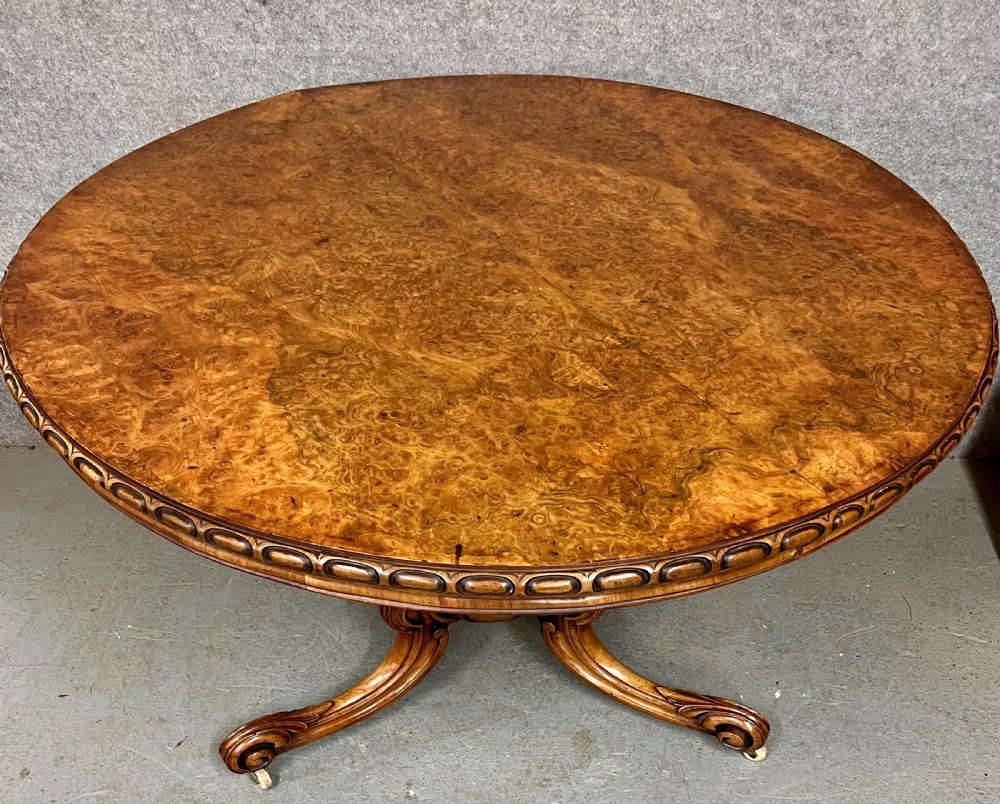 A circular Victorian centre table featuring a figured burr walnut top with carved egg-and-dart edge moulding, supported by a carved central pedestal with four splayed legs on ceramic castors. View VI