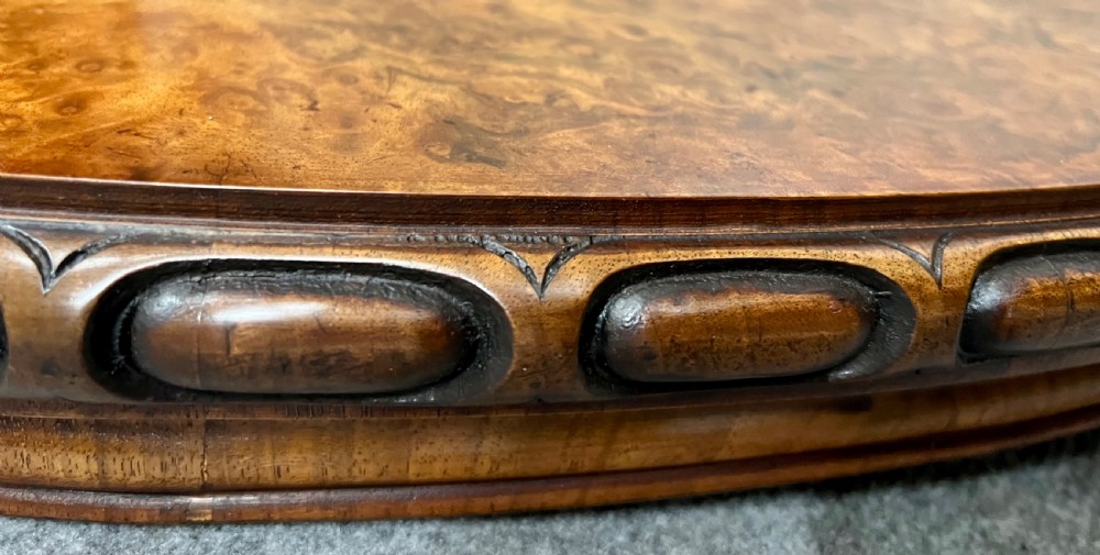 A circular Victorian centre table featuring a figured burr walnut top with carved egg-and-dart edge moulding, supported by a carved central pedestal with four splayed legs on ceramic castors. View IX