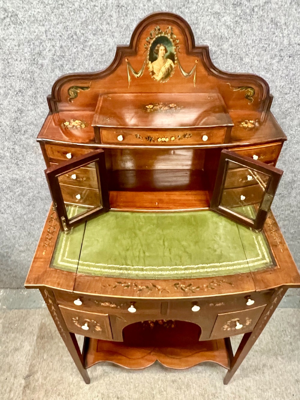 An Edwardian bonheur du jour desk crafted from polished dark wood, featuring a green leather writing surface, painted floral details, a upper superstructure with a painted portrait panel, small drawers, and a central glazed cabinet. View V