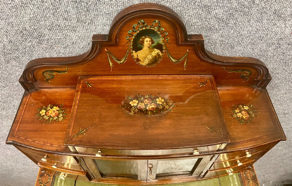 An Edwardian bonheur du jour desk crafted from polished dark wood, featuring a green leather writing surface, painted floral details, a upper superstructure with a painted portrait panel, small drawers, and a central glazed cabinet. View VI
