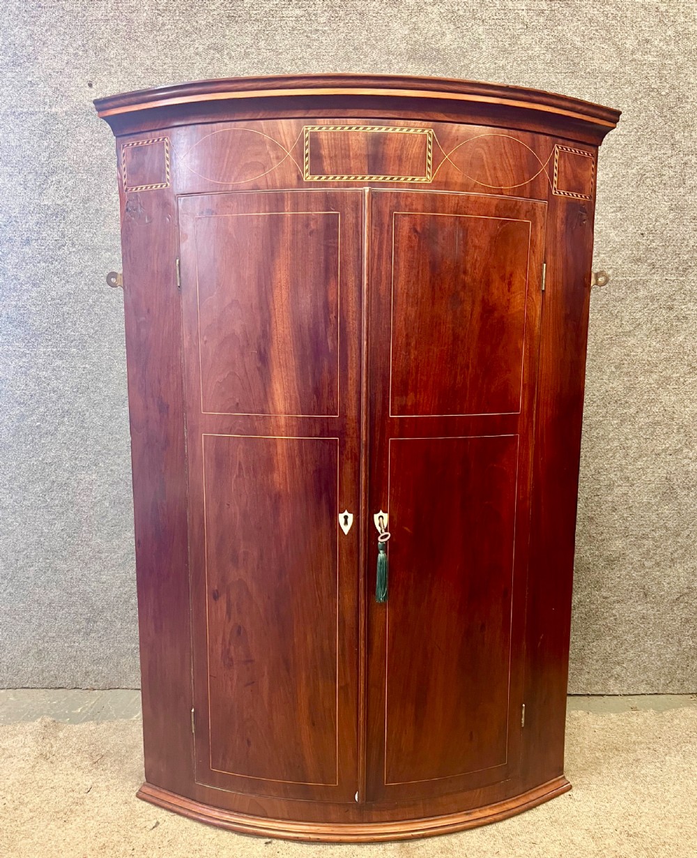 A George III mahogany bow-fronted hanging corner cabinet featuring double doors with fine stringing inlay and shield keyhole escutcheons.