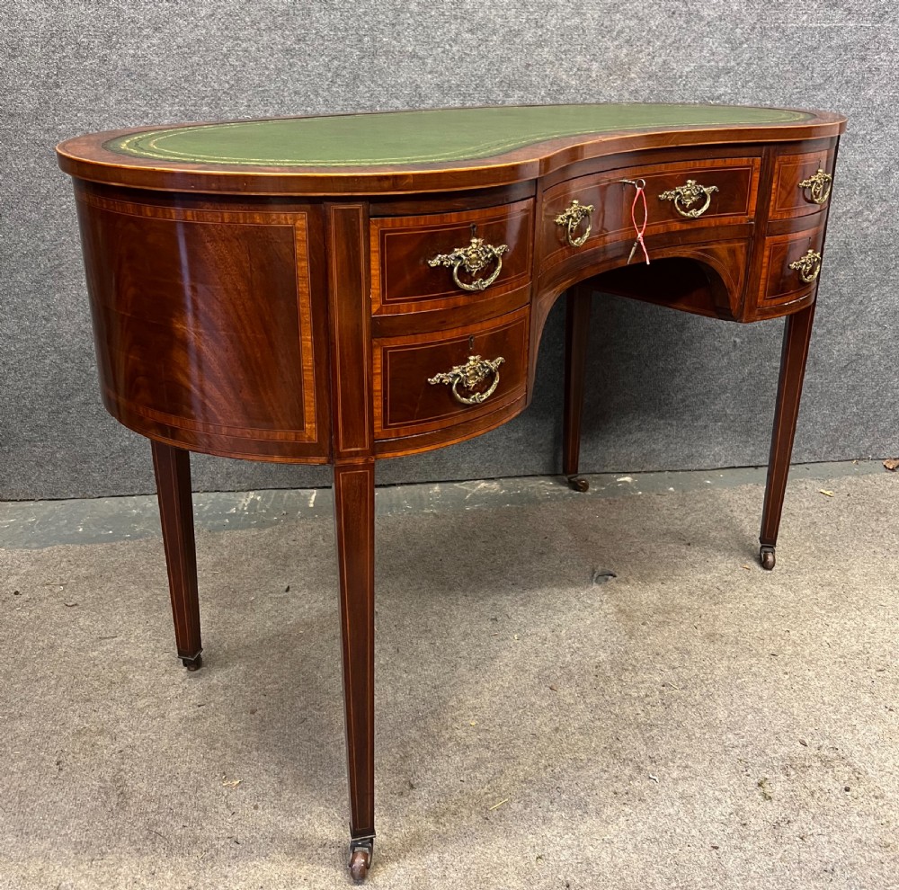 An Edwardian mahogany kidney-shaped desk with a green leather insert top, satinwood inlay, five drawers, and tapered legs on brass castors. View V