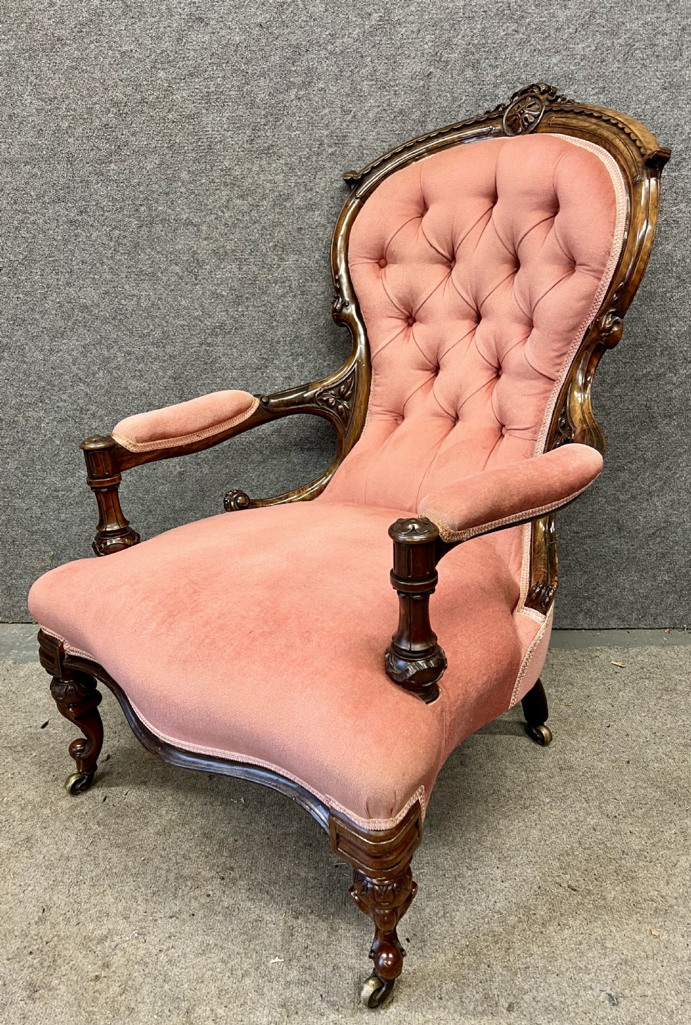 A Victorian open armchair crafted from carved dark walnut with button-backed rose pink velvet upholstery and brass castors.